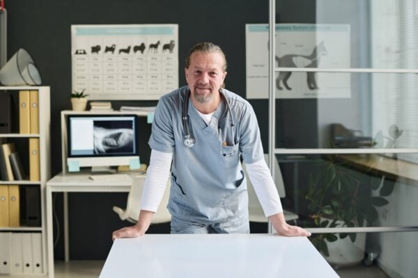 Middle-aged veterinarian leaning on examination table at vet office in clinic looking at camera